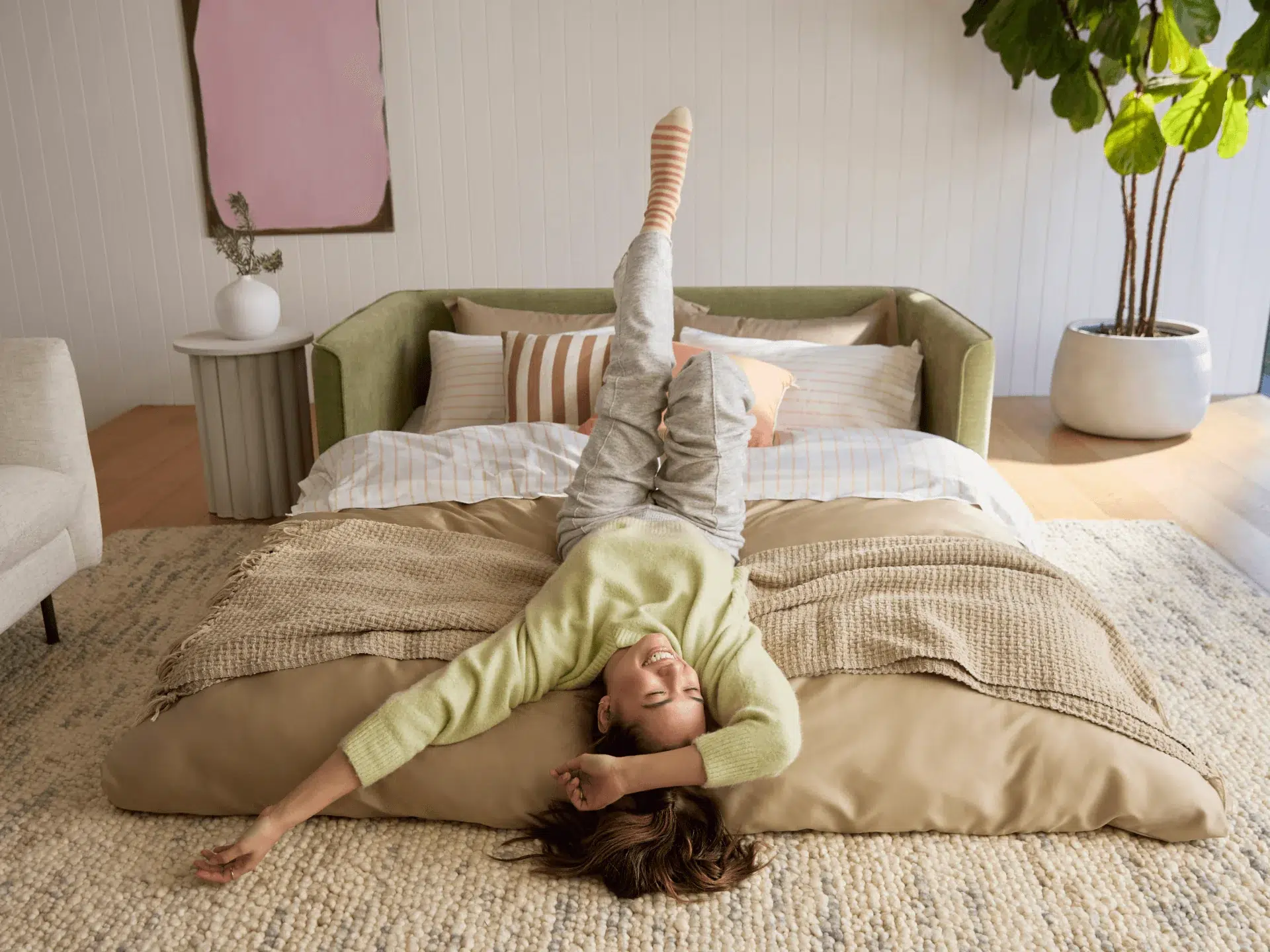 A woman lies on a bed with her arm stretched out and one leg raised, wearing casual clothes and striped socks in a bright, modern room.