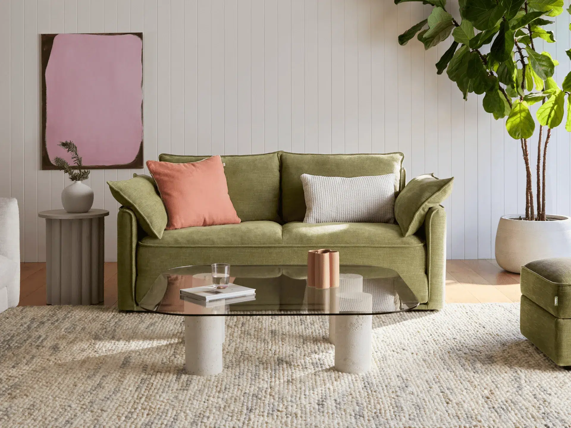 A green sofa with pink and gray cushions sits in a bright living room, featuring a glass coffee table, textured rug, potted plant, and abstract wall art.