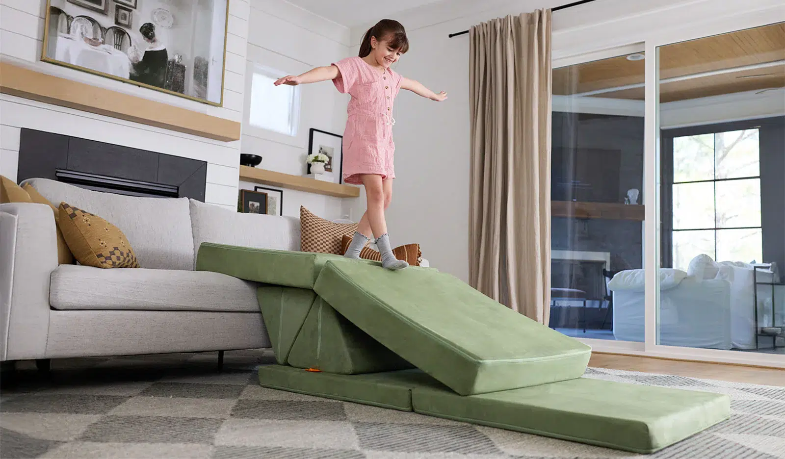 A young girl in a pink romper stands with arms outstretched on a green foam playset positioned on a living room couch, with sliding glass doors and neutral decor in the background.