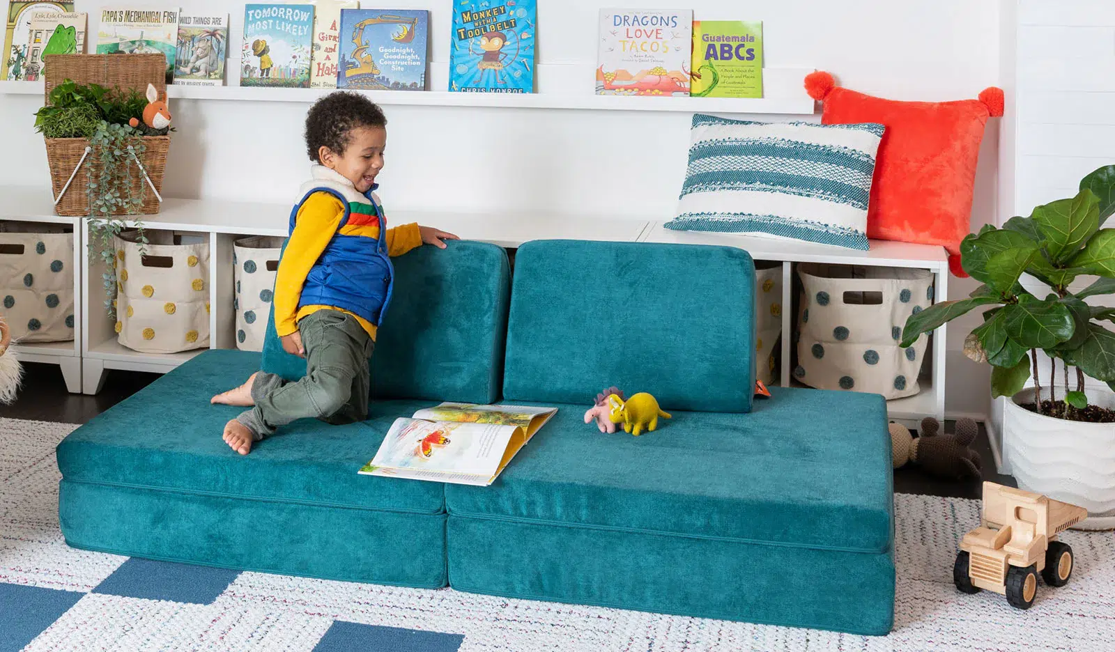 A young child sits and plays on a teal modular couch in a playroom with books, toys, and storage baskets in the background.