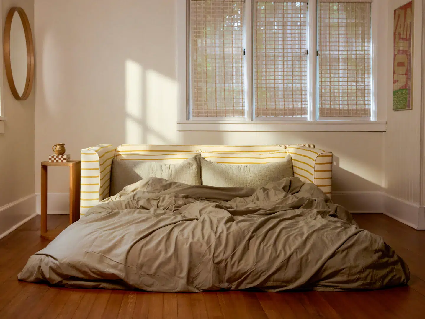 A bed with rumpled beige bedding sits on a wooden floor in front of a striped yellow sofa, with a small side table and three windows with blinds in the background.