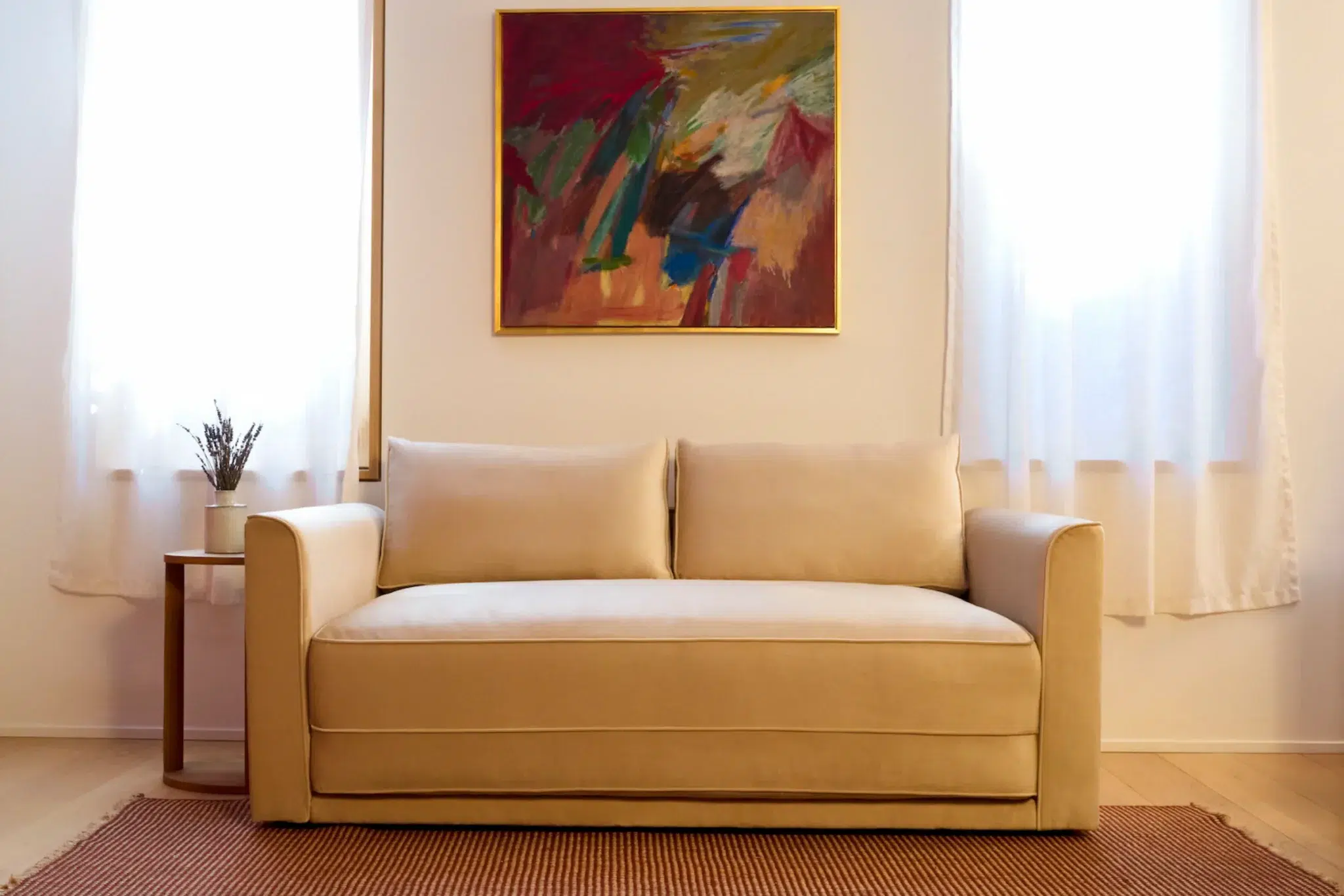 A beige sofa sits on a brown rug between two windows with white curtains, a small side table with a vase, and a colorful abstract painting on the wall above.