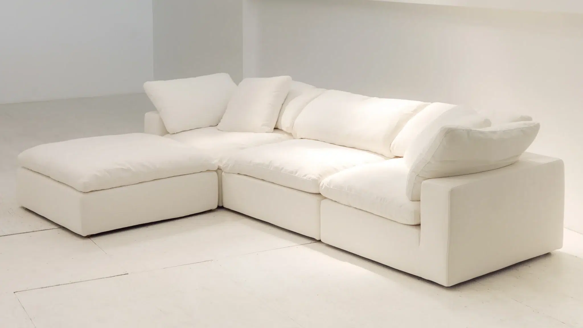 A modern, white, L-shaped sectional sofa with plush cushions and a chaise lounge, placed in a minimalist, light-colored room.