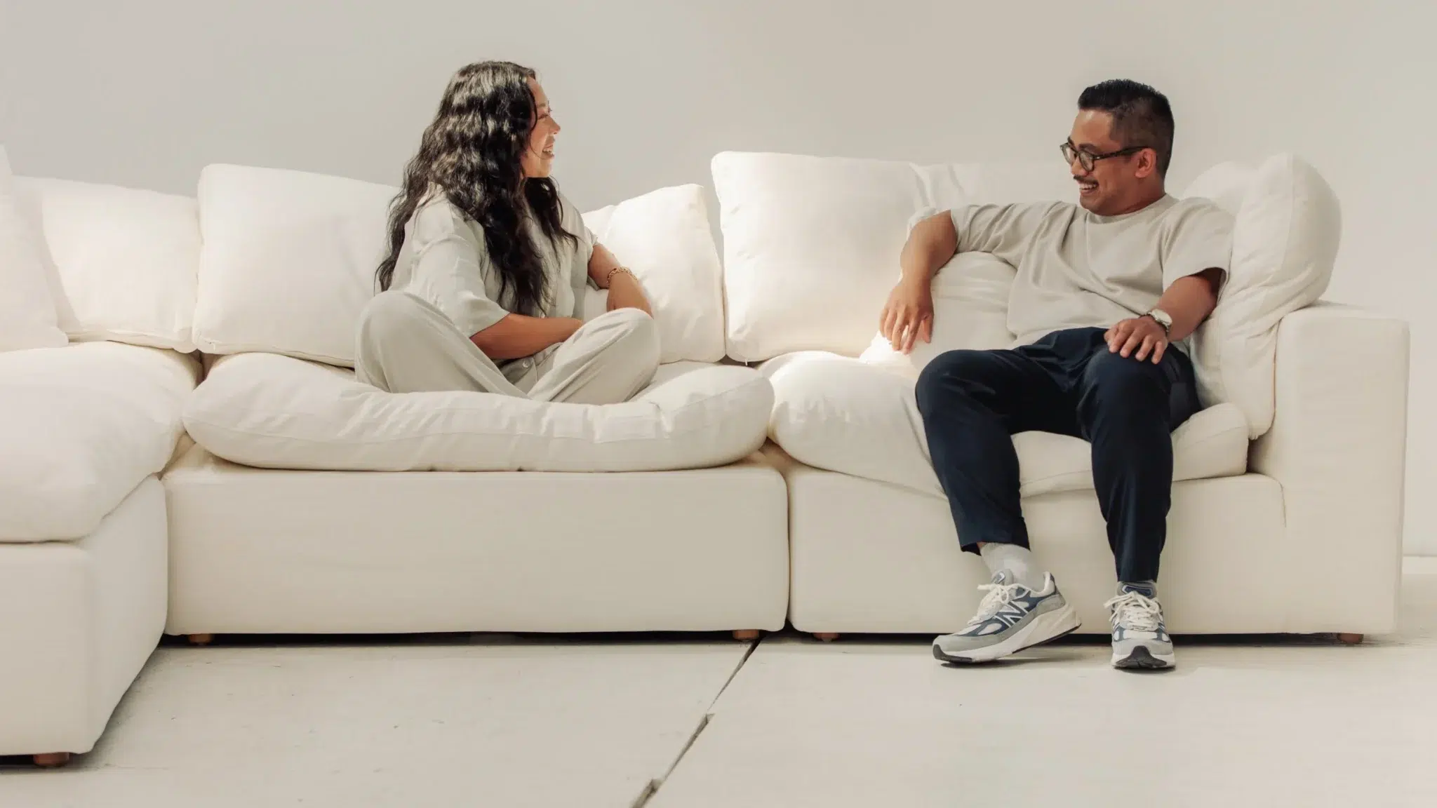 Two people sit on a white sofa facing each other, engaged in conversation, in a brightly lit room with a minimalistic style.