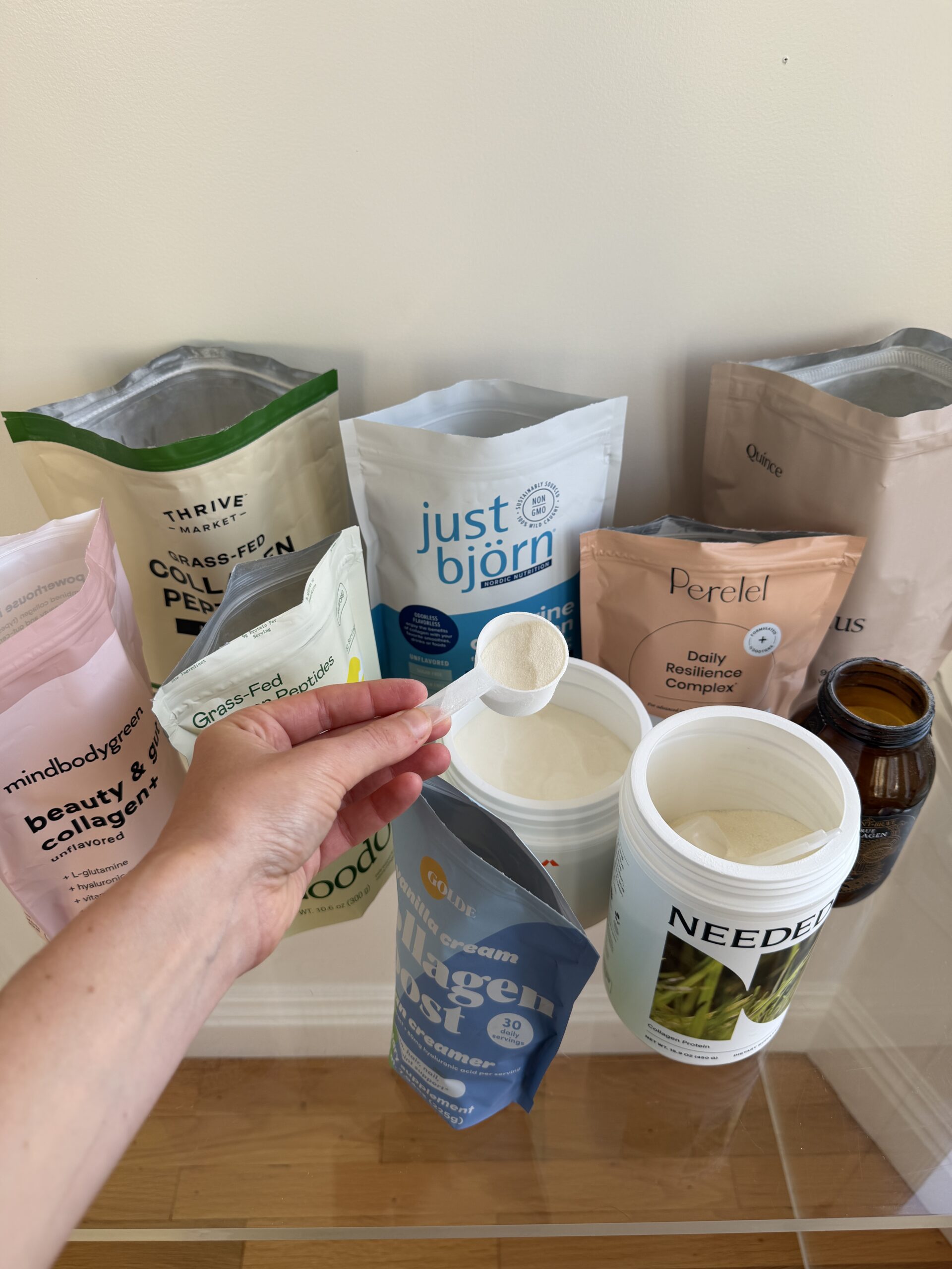 A hand holds a scoop of white powder over an open container, surrounded by various collagen and protein supplement packages on a wooden surface.