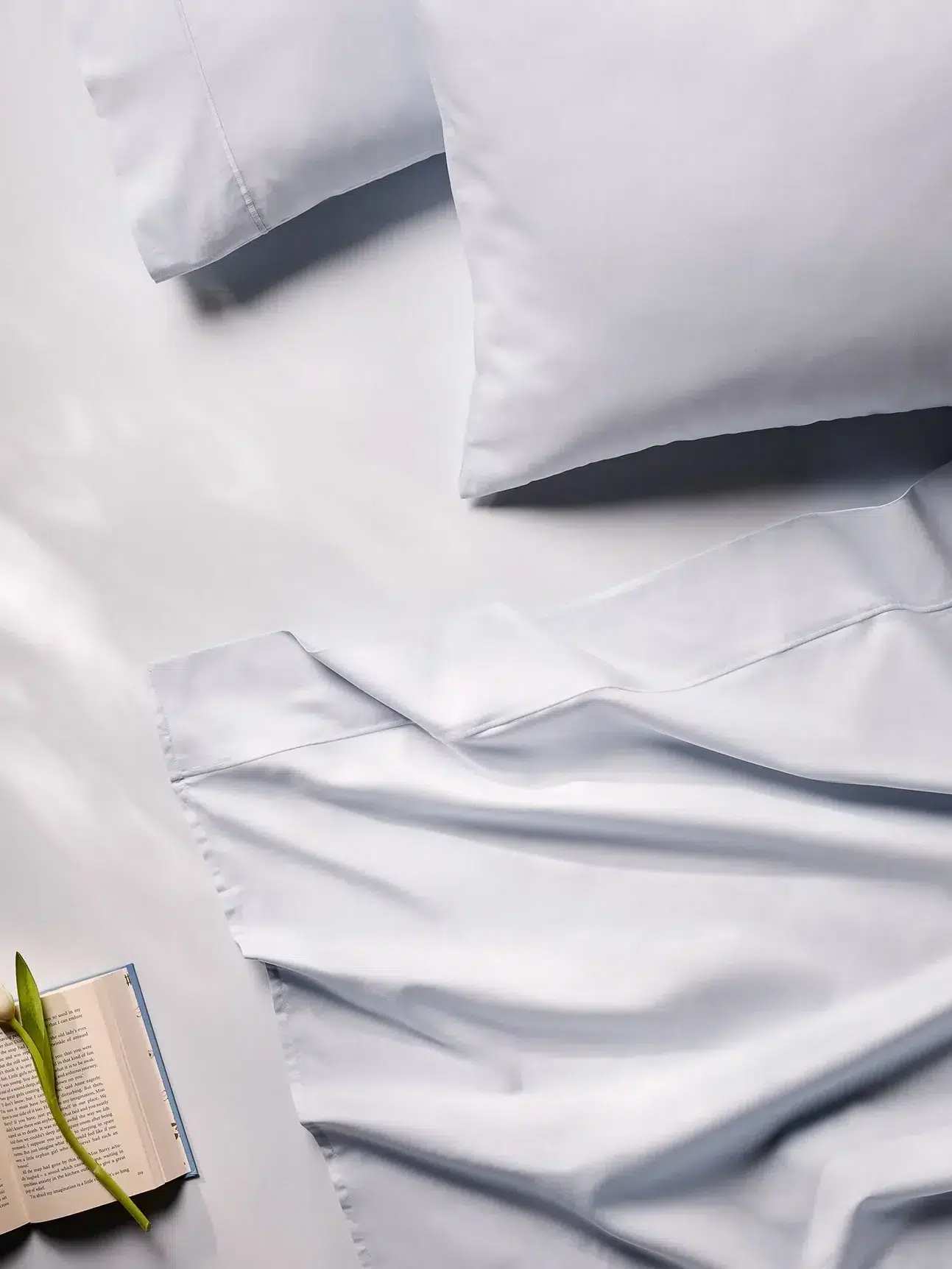 A neatly made bed with light gray sheets and two pillows, next to an open book and a single white tulip on a flat surface.