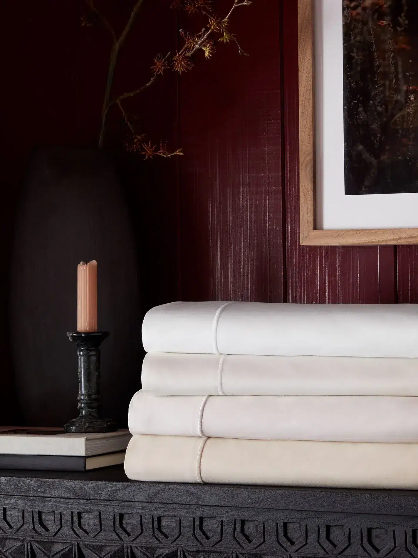 Four folded cream and white bedsheets are stacked on a dark carved table next to a candle, a vase with branches, and a framed picture on a dark red paneled wall.