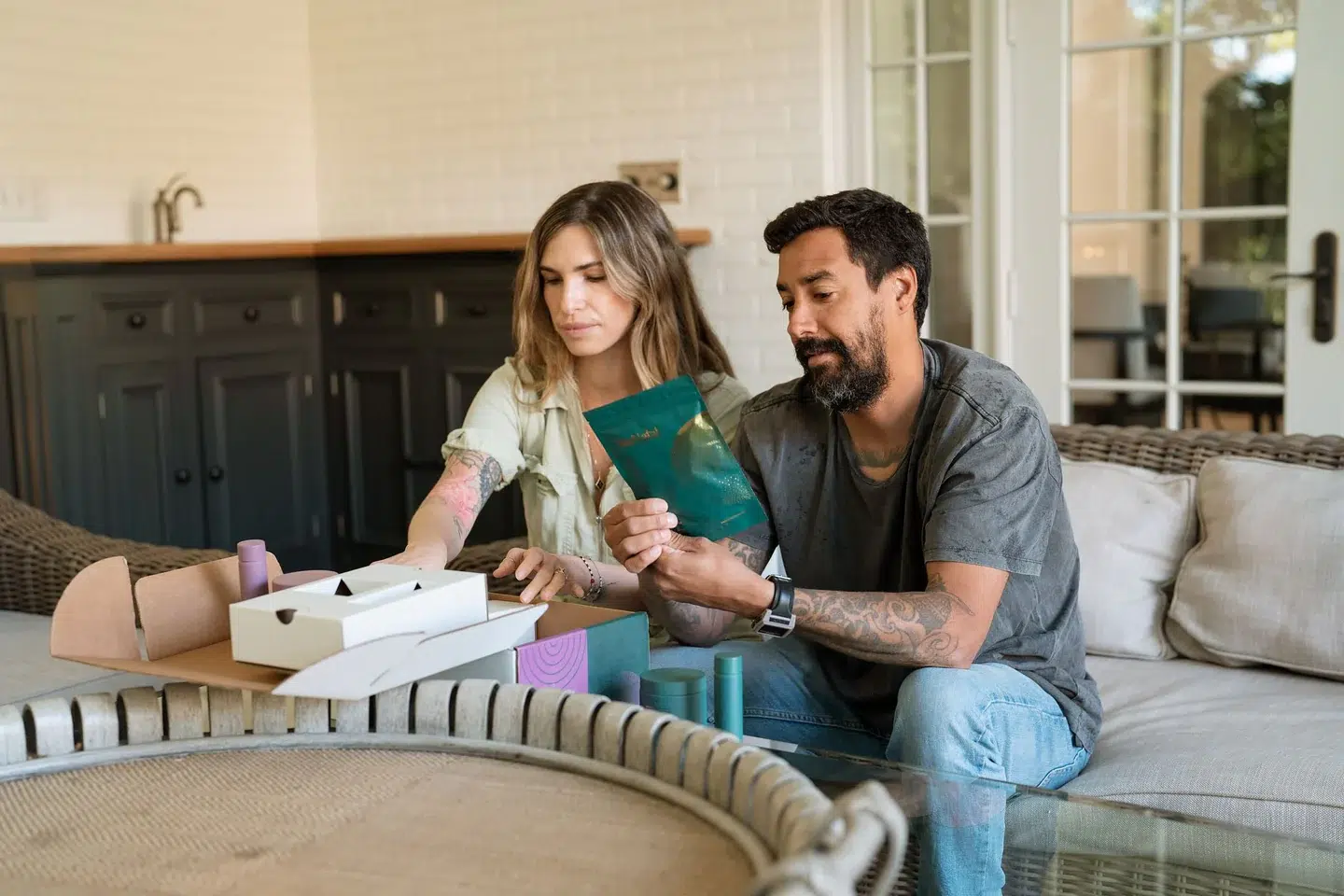 A woman and a man sit on a couch examining packaging and products from boxes on a table in a home setting.
