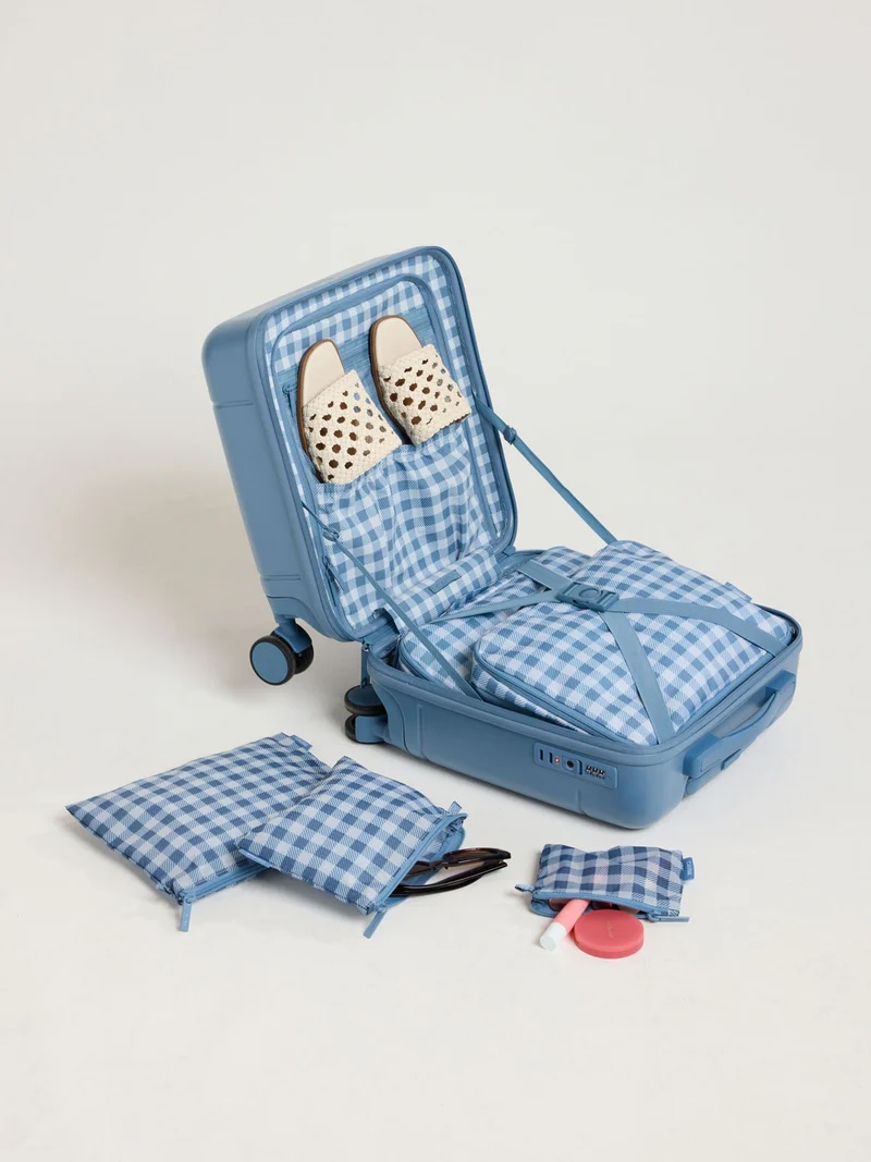 A blue gingham-pattern suitcase is open, showing matching packing cubes inside, a pair of slip-on sandals, and toiletries. Additional packing cubes and items are placed beside the suitcase.