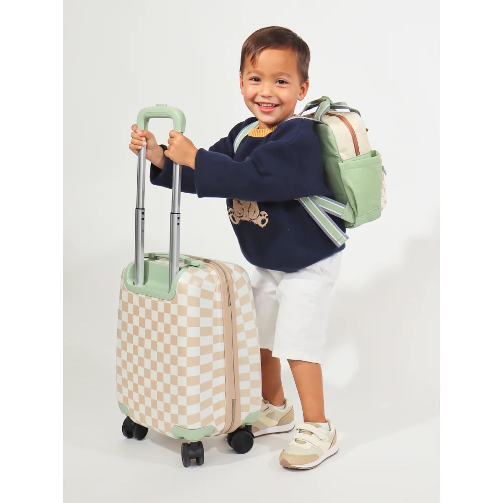 A young child wearing a navy sweater, white shorts, and sneakers stands smiling with a checkered suitcase and a matching backpack.