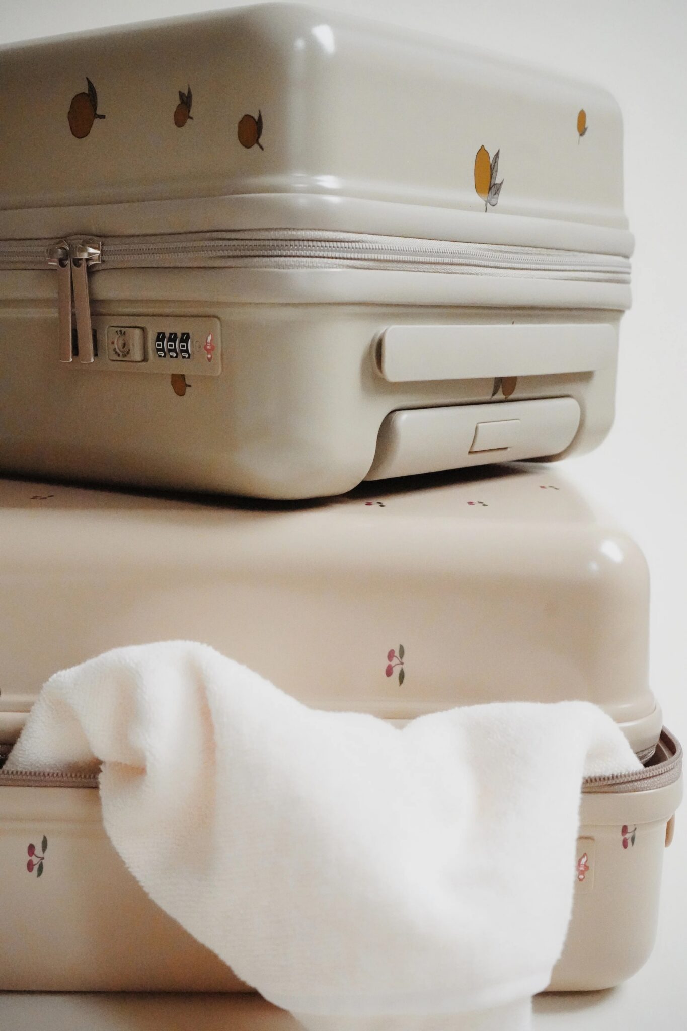 Two beige hard-shell suitcases with small fruit designs are stacked on each other, with a white towel draped over the lower suitcase.