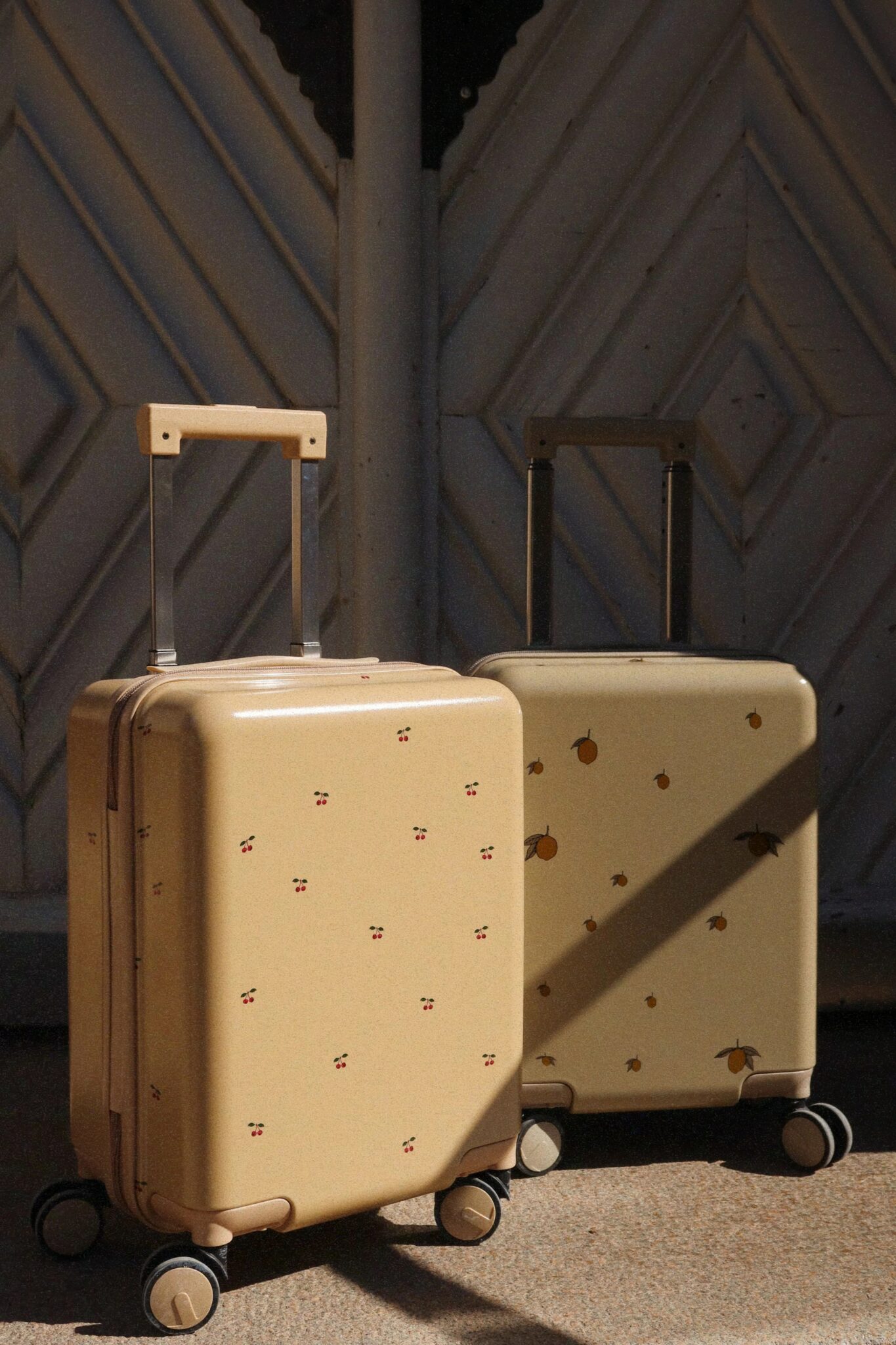 Two beige hard-shell suitcases with small decorative prints stand side by side on a textured floor in sunlight, positioned in front of a patterned wall.