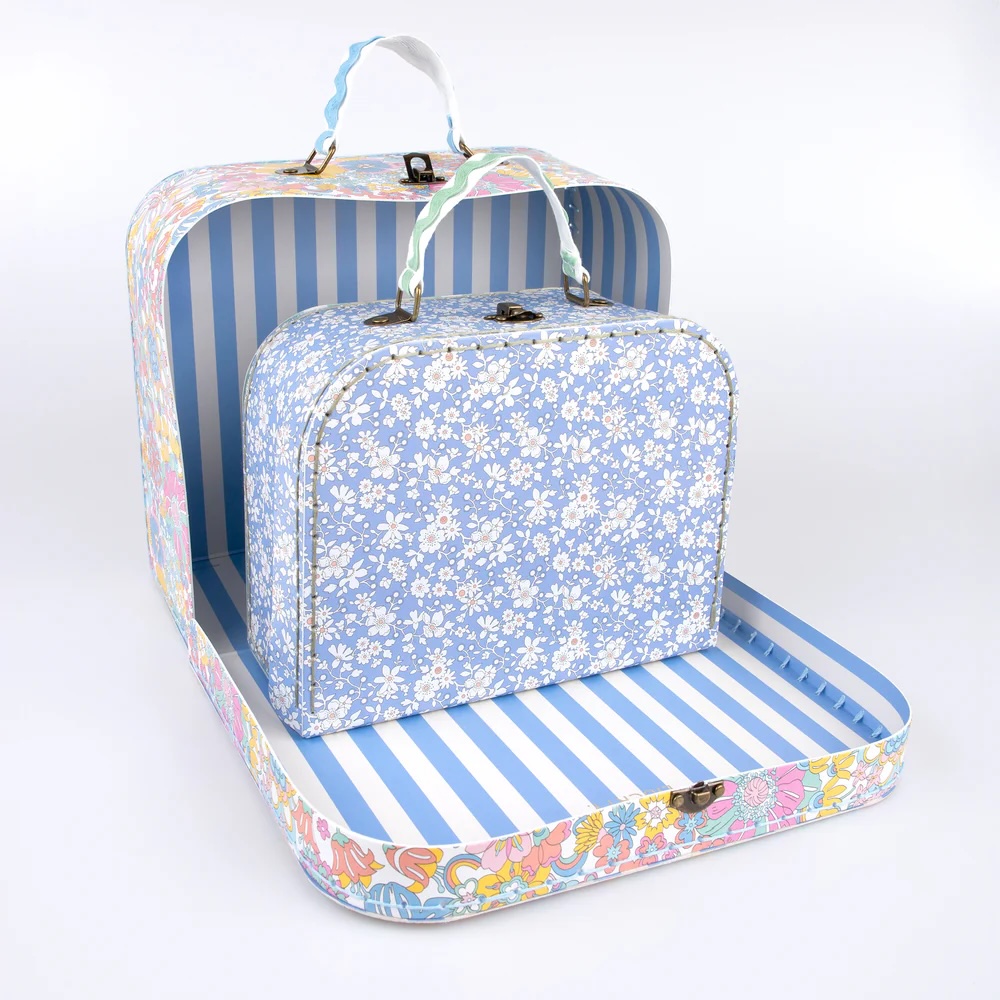 Two decorative suitcases: a larger one with a floral and striped interior, holding a smaller, blue suitcase with a white floral pattern. Both have handles and clasps.