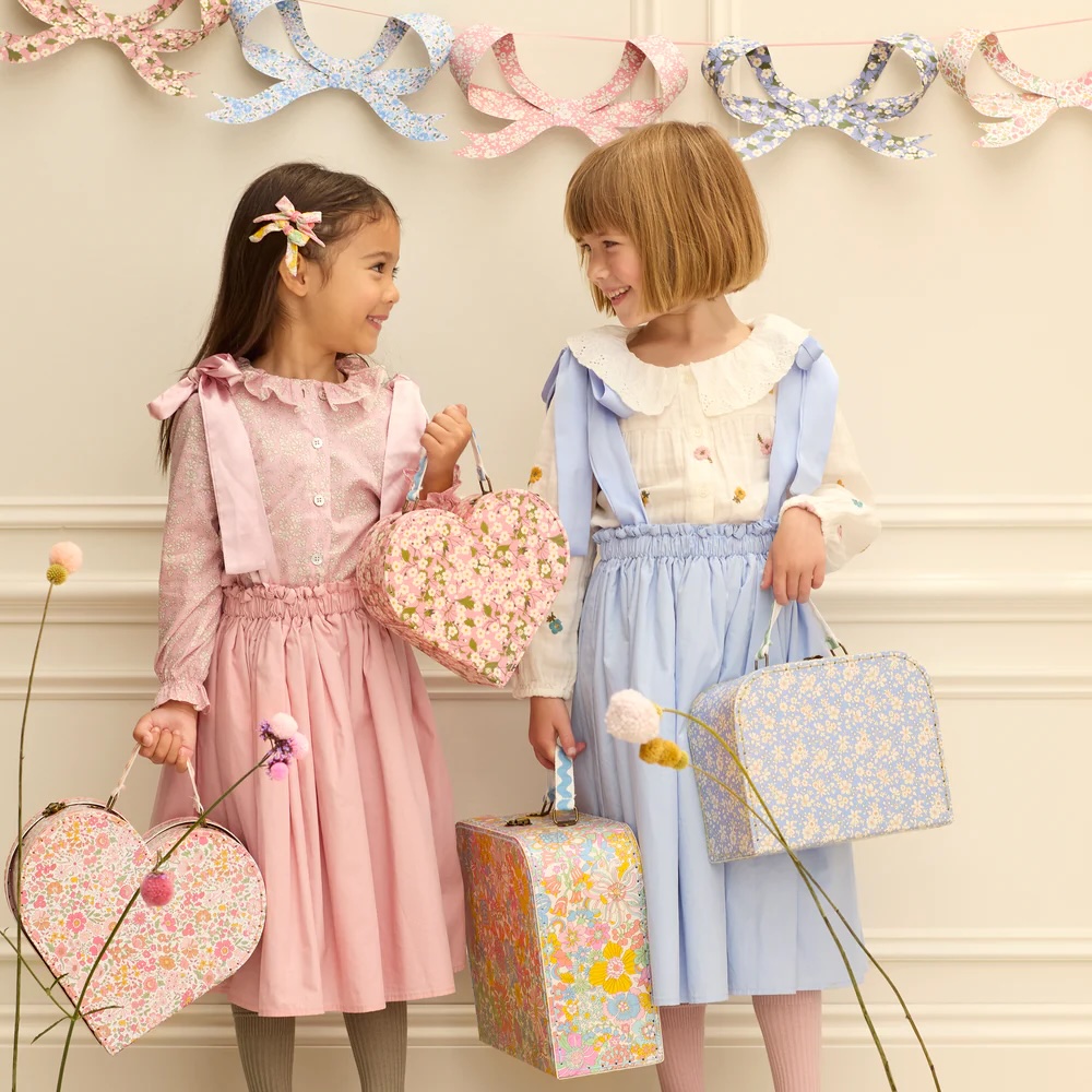 Two young girls wearing pastel dresses and holding floral suitcases stand in front of a white wall decorated with floral garlands, smiling at each other.