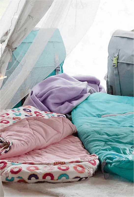 A close-up of colorful sleeping bags and blankets arranged inside a tent, with backpacks and soft lighting in the background.