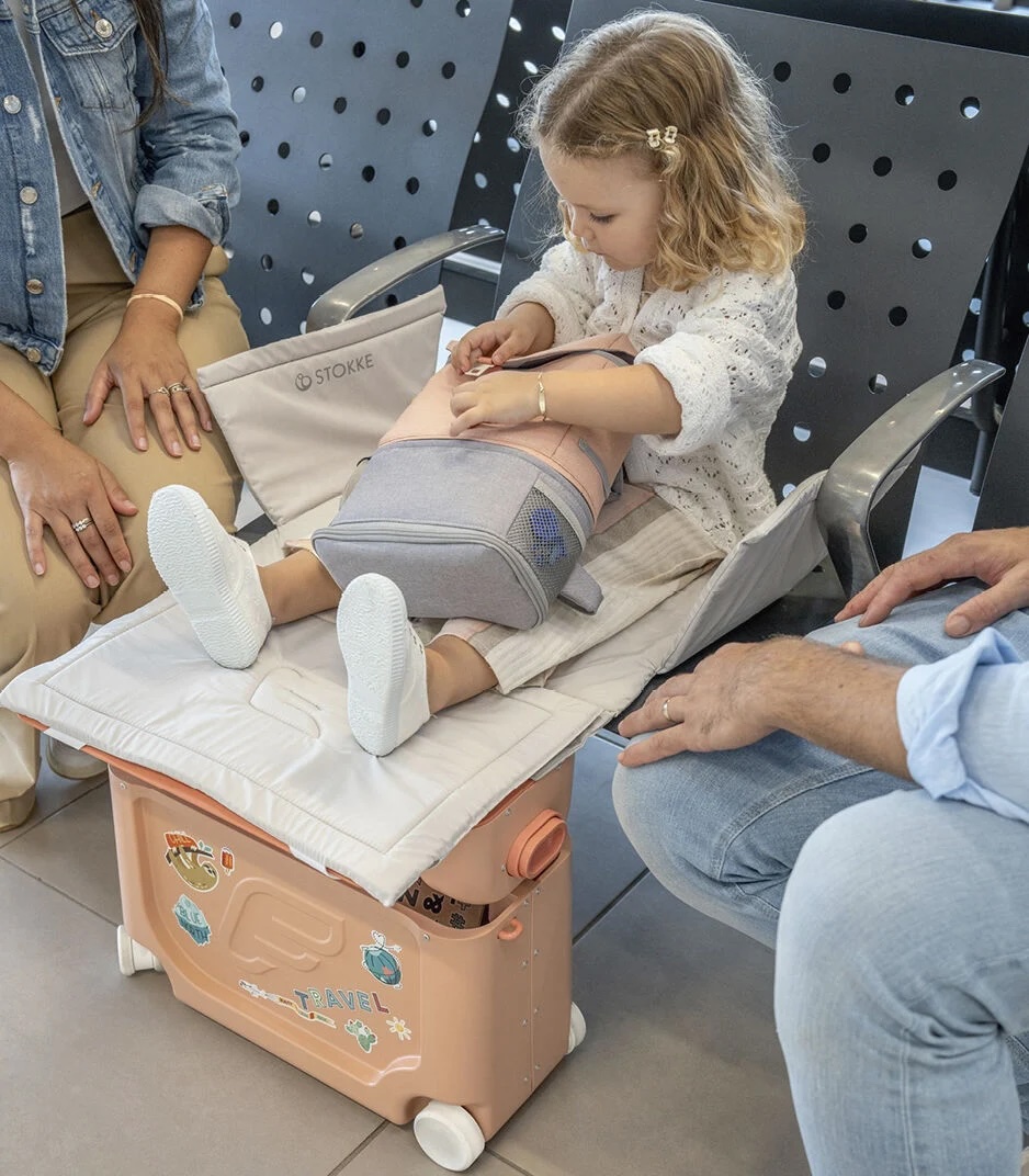 A young child sits on an extended travel seat, using a suitcase as a footrest, while two adults sit nearby in an airport waiting area.