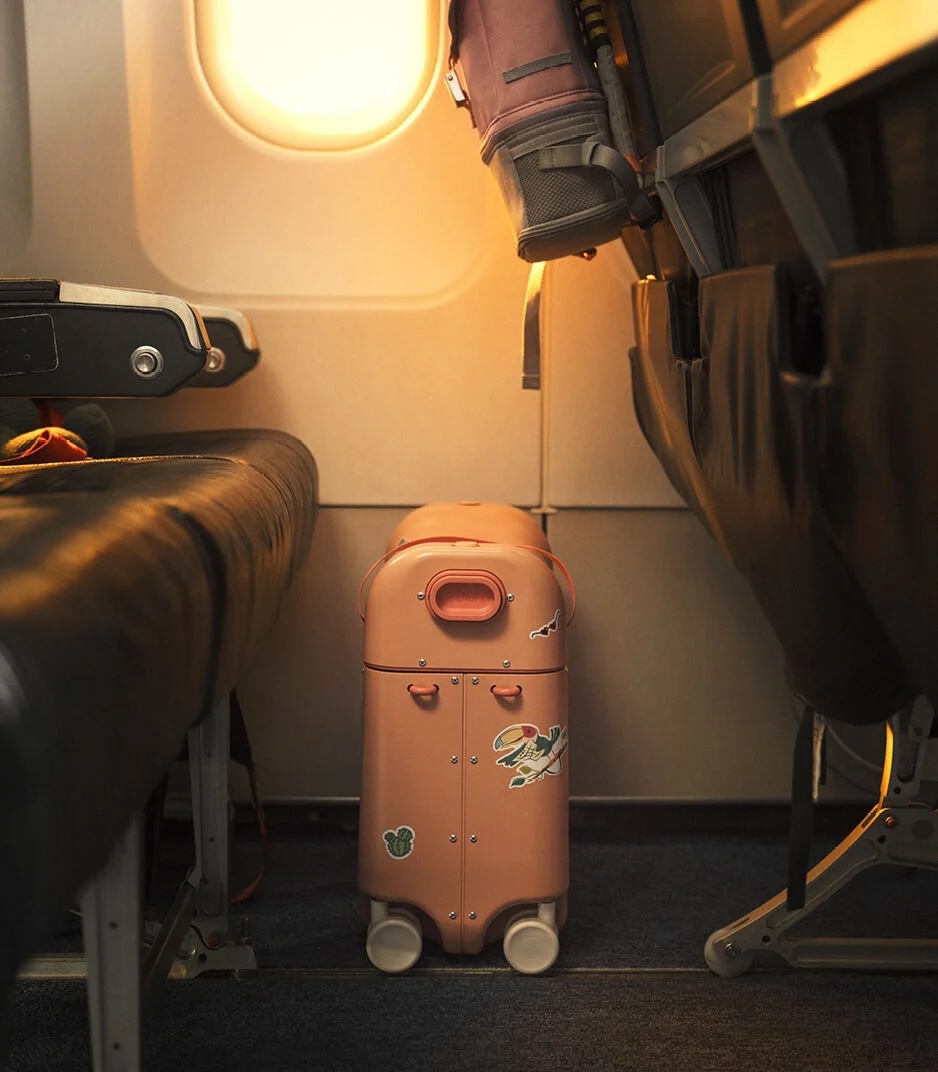 Pink suitcase with stickers is placed under an airplane seat next to a window, with light shining in from outside.