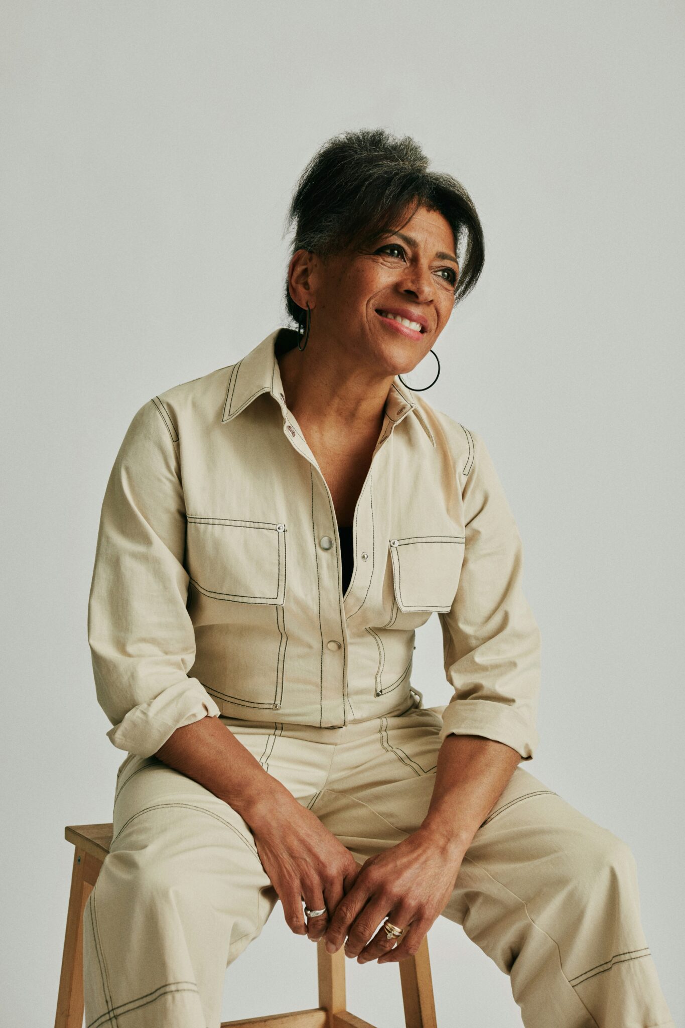 A woman with gray hair, wearing a beige utility jumpsuit, sits on a wooden stool against a plain light background, smiling and looking slightly to the side.
