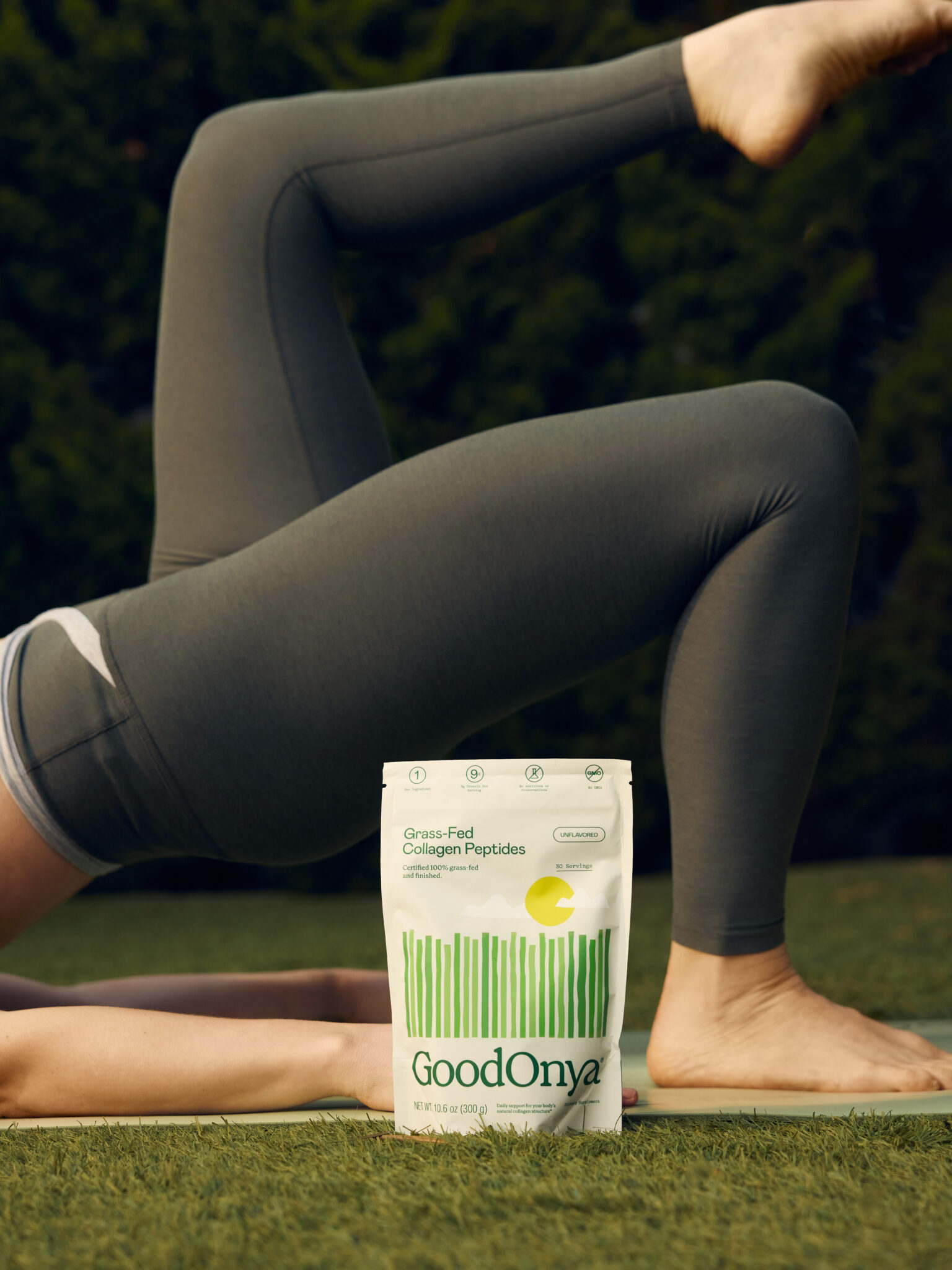 A person in dark leggings does a yoga pose on a mat outdoors, with a GoodOnya Grass-Fed Collagen Peptides pouch placed in front of them on the grass.