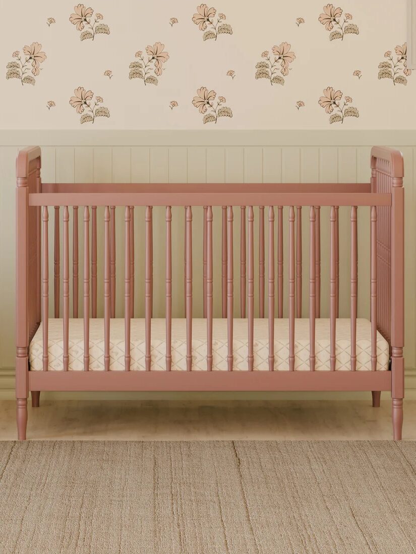 A pink wooden crib with a beige patterned mattress is placed against a wall with floral wallpaper and two windows, on a beige rug.