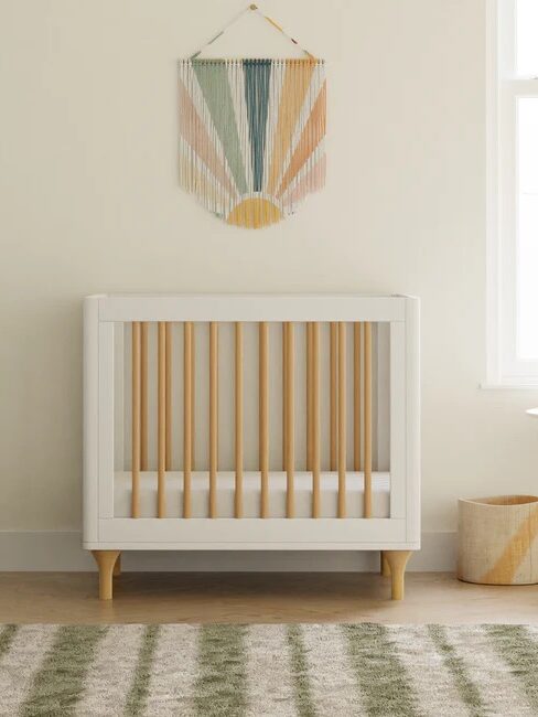 A modern nursery with a white crib featuring wooden slats, a white dresser, a striped rug, and a colorful wall hanging above the crib.