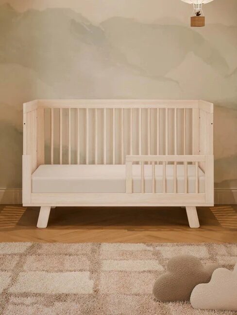 A light wood toddler bed with a safety rail is placed against a wall with soft green and beige tones, on a beige rug with cloud-shaped pillows nearby.