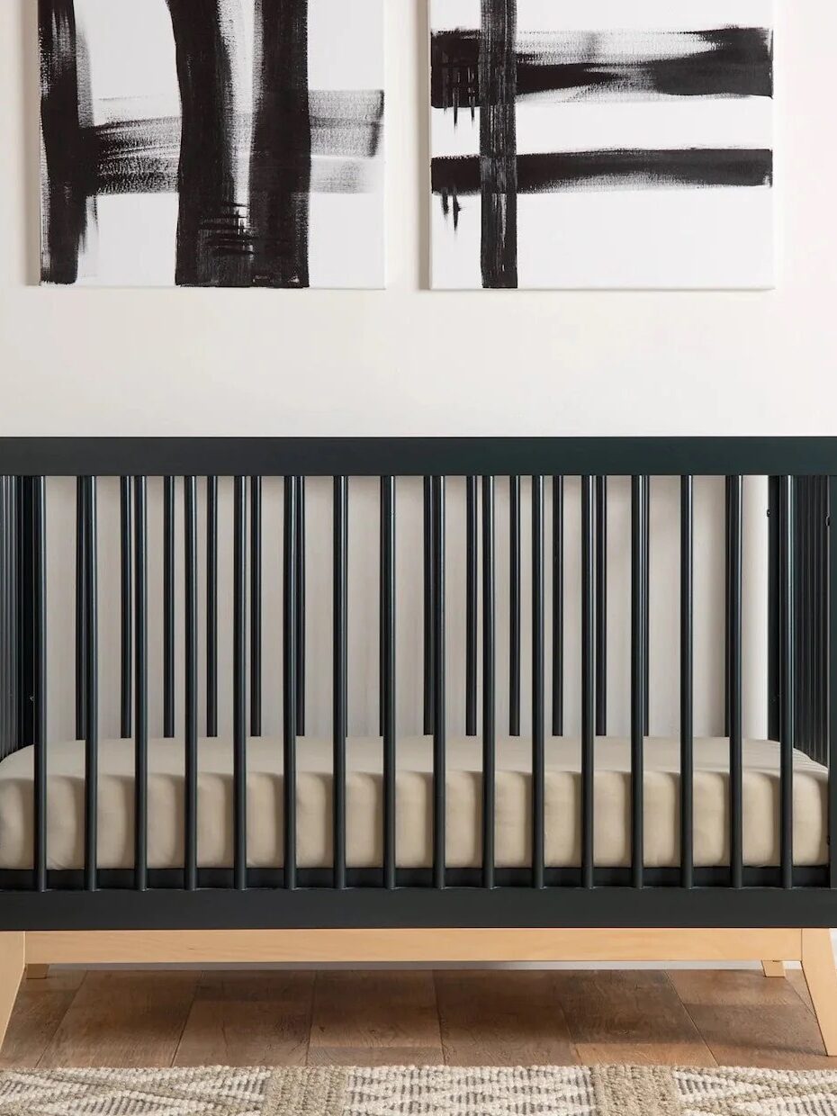 A modern black crib with light wood legs and a mattress, placed against a wall with abstract black and white artwork above it, on a wooden floor with a patterned rug.