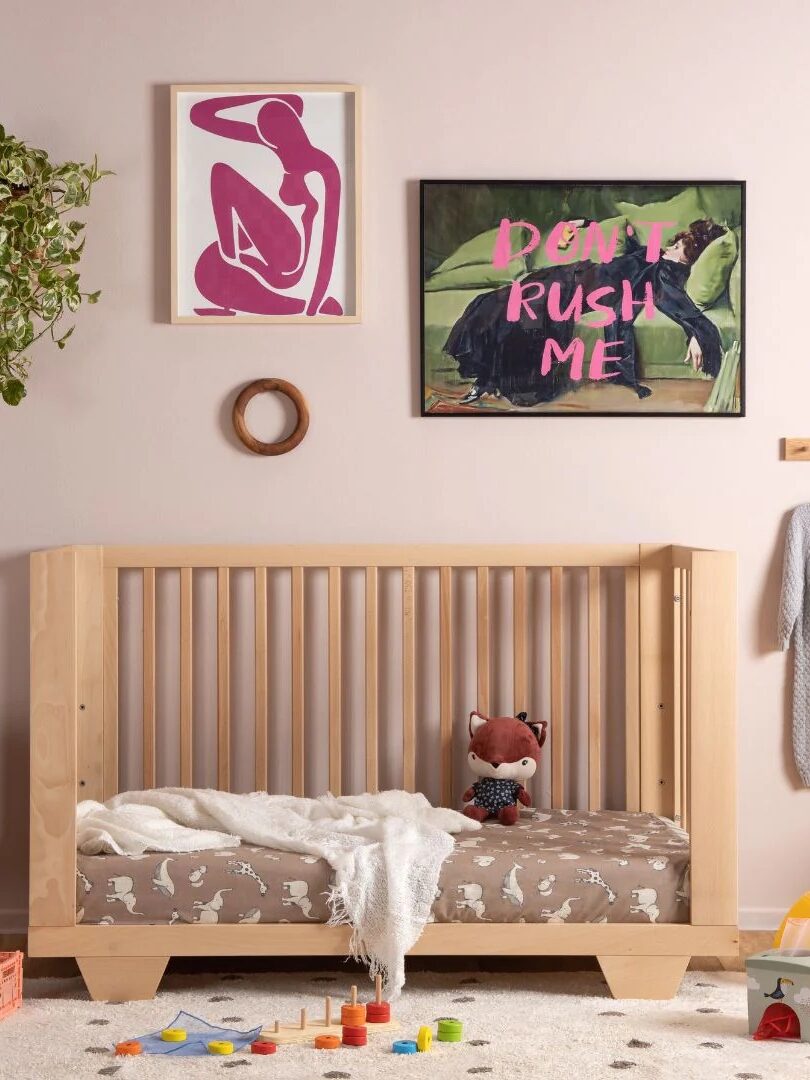 A wooden crib with animal-patterned bedding, a stuffed fox, and a white blanket sits against a pink wall with two framed artworks and a hanging plant above it; toys are scattered on the floor.