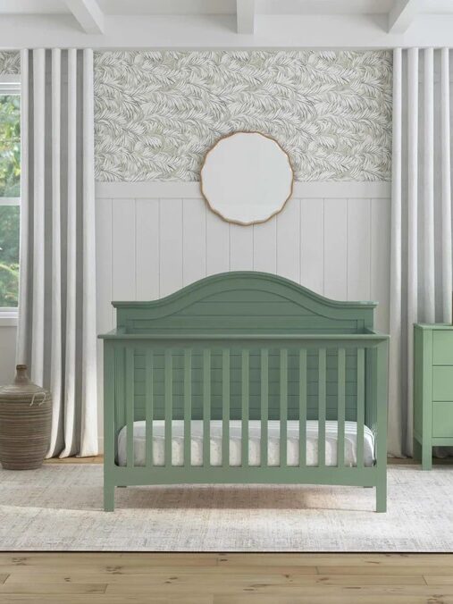 A green crib is centered in a nursery with patterned wallpaper, white wainscoting, matching green dresser, round mirror, floor lamp, and large windows with white curtains.