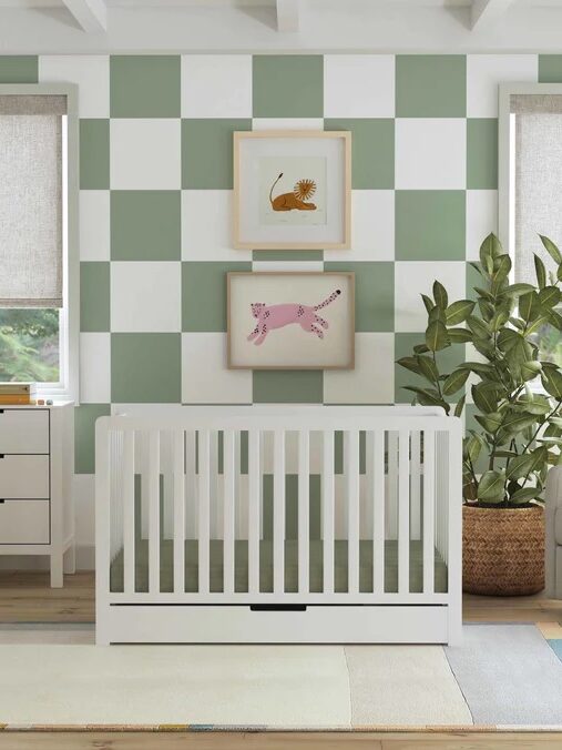A modern nursery with a white crib, a checkered green and white accent wall, framed animal art, a dresser, a potted plant, and two windows with light-colored blinds.