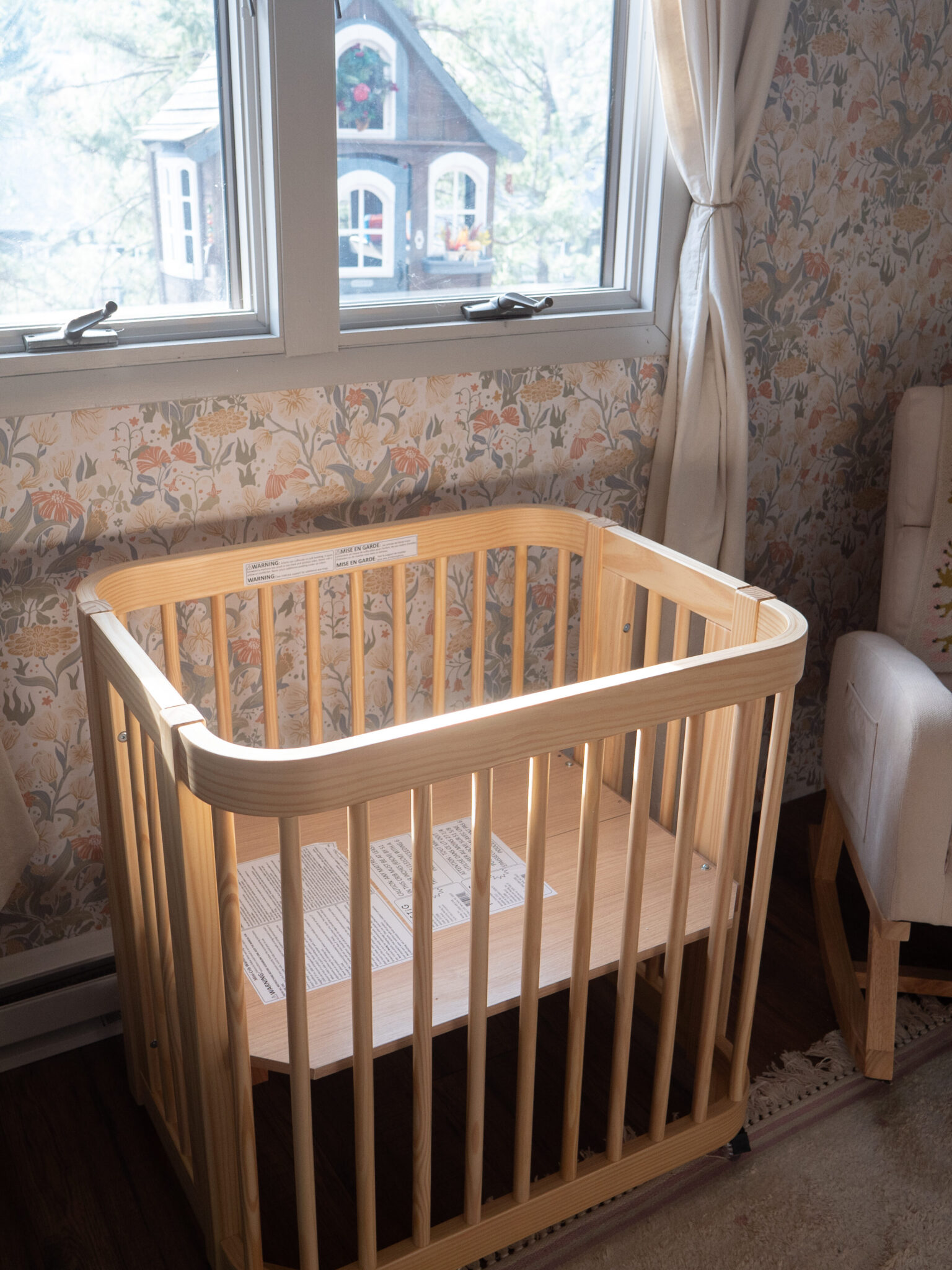 A wooden crib sits near a window in a softly lit nursery with floral wallpaper, a white armchair, and a small green stool.