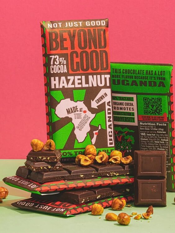 A bar of Beyond Good hazelnut chocolate stands upright with pieces of chocolate and scattered hazelnuts in front, against a pink and green background.