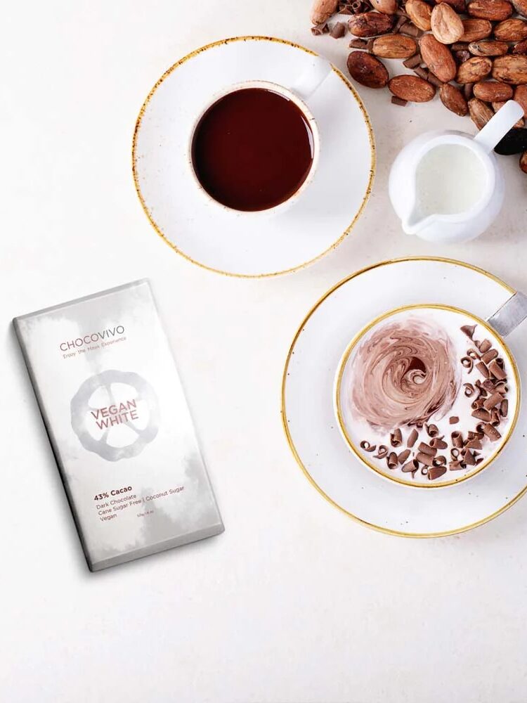 A cup of hot chocolate with chocolate shavings, a cup of chocolate drink, a small creamer, cocoa beans, and a Chocovivo Vegan White chocolate bar on a white surface.