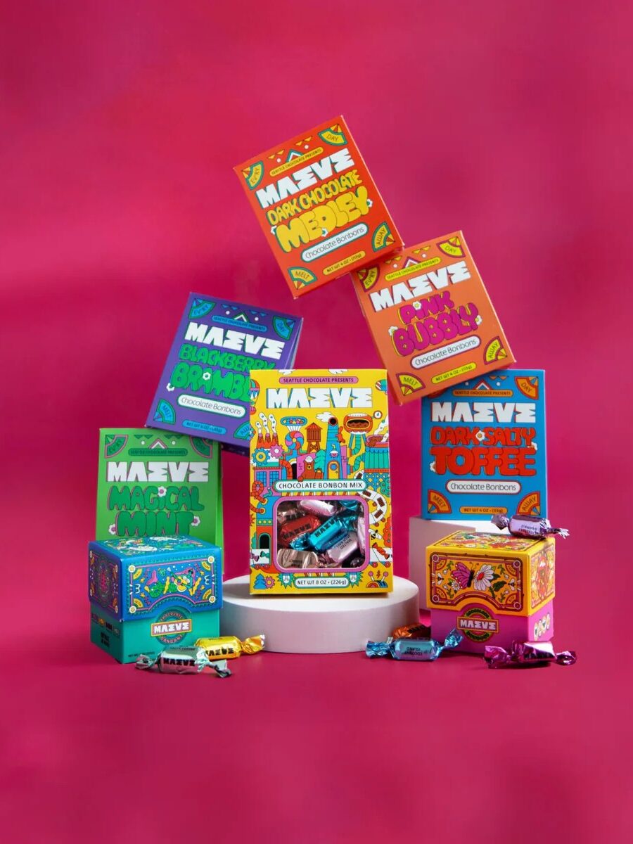 A variety of colorful MAEVE candy boxes and wrapped candies are arranged on a white platform against a bright pink background.