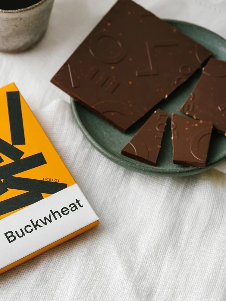 A yellow and white chocolate bar wrapper labeled "Buckwheat" beside a green plate with pieces of dark chocolate and a ceramic cup, all on a white cloth surface.
