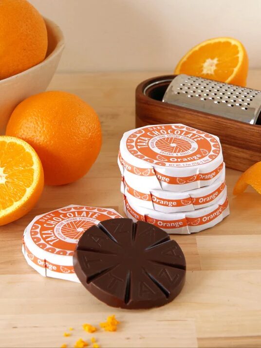 A stack of orange-themed chocolate discs sits on a wooden surface near fresh oranges, orange peel, a grater, and a bowl of whole oranges. One chocolate disc is broken into segments.