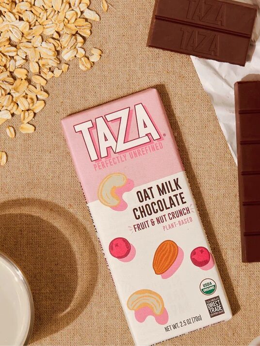 Taza Oat Milk Chocolate bar on a table with chocolate pieces, oats, a glass of oat milk, and a small bowl of dried fruit and nuts.