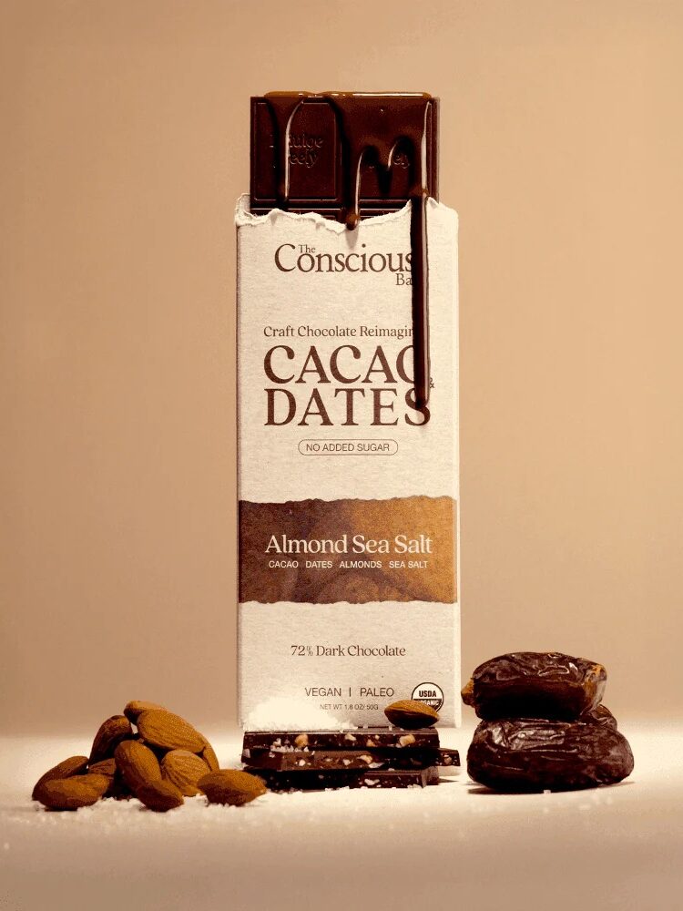 A chocolate bar labeled “Cacao Dates Almond Sea Salt” stands upright with chocolate melting on top, surrounded by whole almonds and dates against a neutral background.