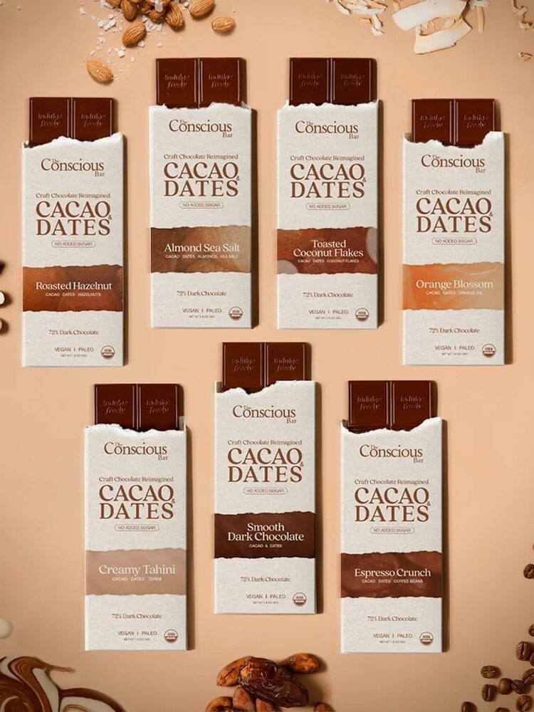 Seven Conscious Cacao Dates chocolate bars are arranged on a beige surface with scattered almonds, chocolate pieces, orange slices, and nuts around them.