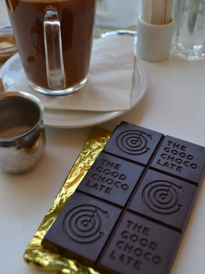 A glass mug of coffee sits on a plate beside a bar of dark chocolate labeled "The Good Chocolate," partially unwrapped on a gold foil wrapper.