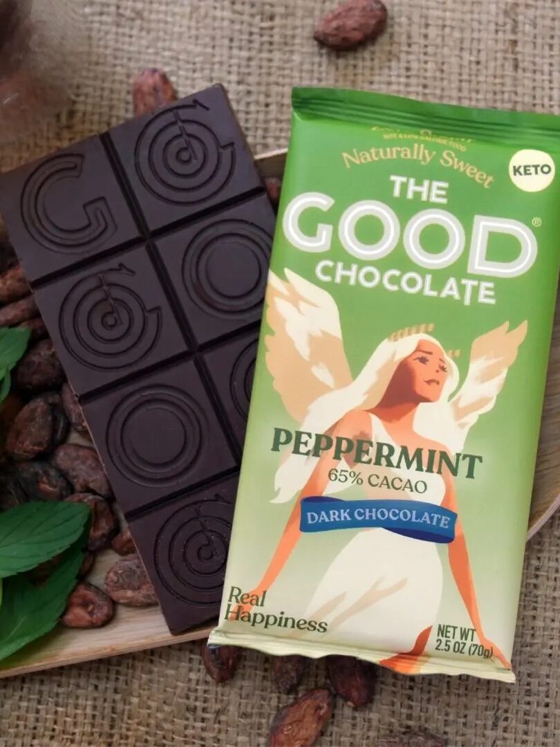 A bar of dark chocolate with geometric patterns sits next to a green package labeled "The Good Chocolate Peppermint 65% Cacao" on a wooden tray with mint leaves and cocoa beans.