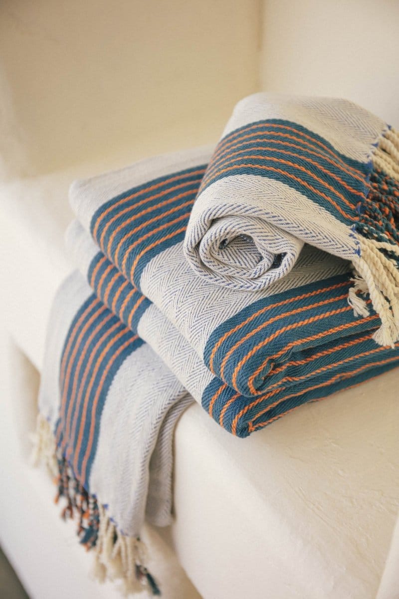 Two folded and one rolled fringed blankets with blue, orange, and cream stripes are stacked on a white surface.