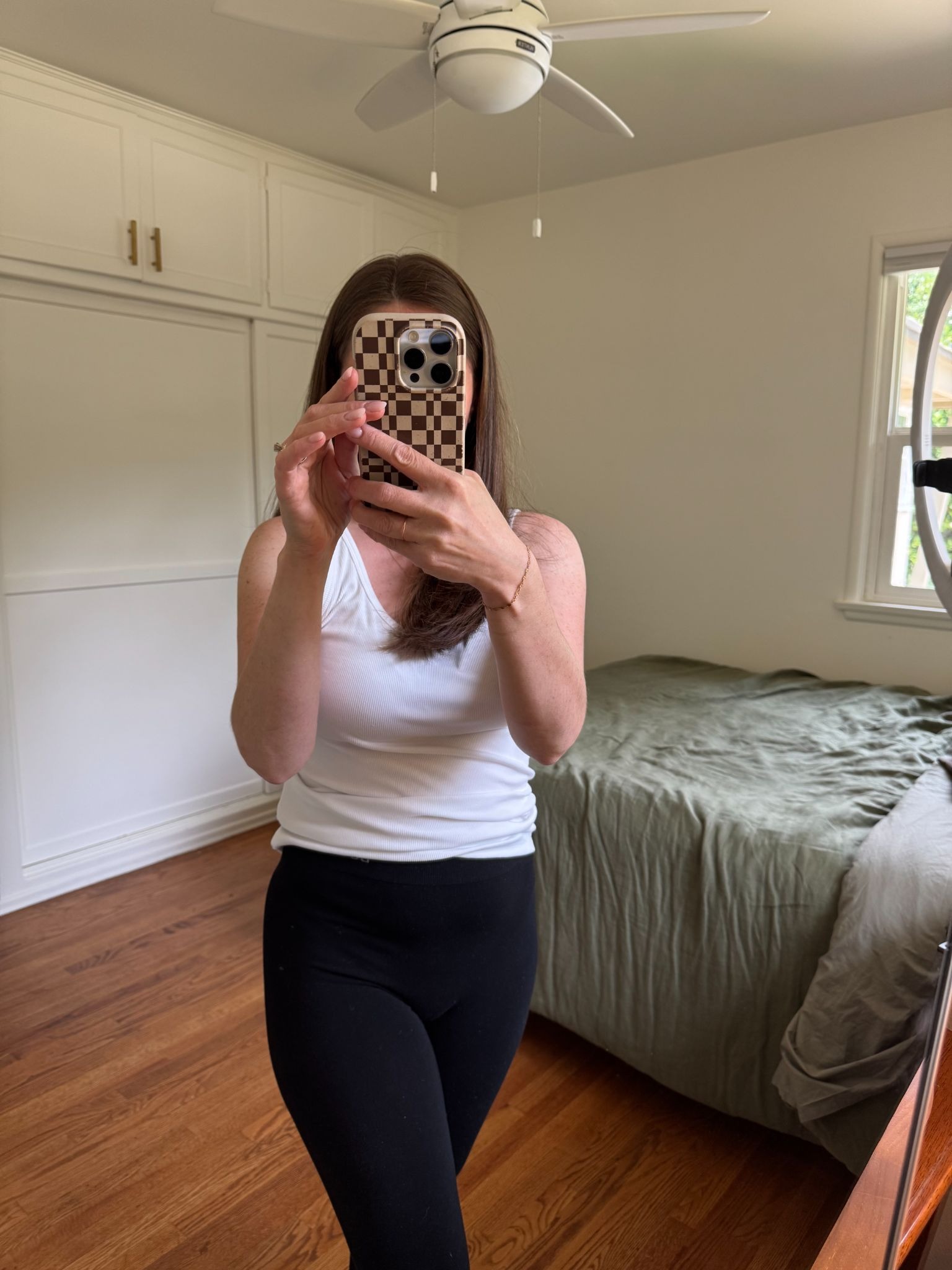 A person takes a mirror selfie in a bedroom, wearing a white tank top and black pants. The room has wooden floors, a made bed, and white walls.