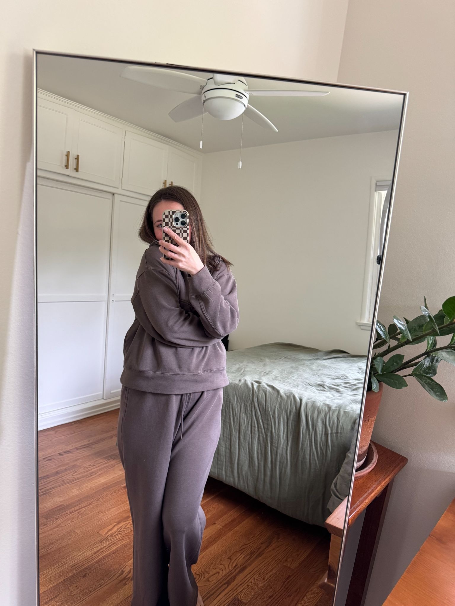 Person taking a mirror selfie in a bedroom, wearing a brown sweatsuit and holding a phone with a patterned case; bed, plant, and ceiling fan visible in the background.
