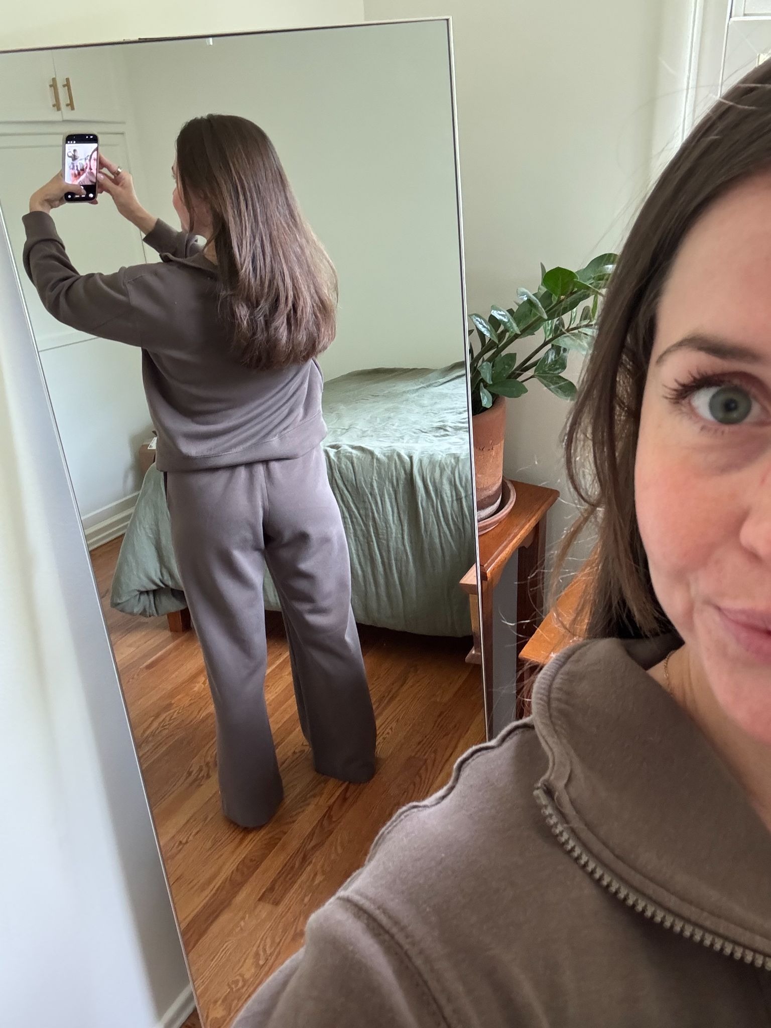 A woman in brown loungewear takes a mirror selfie, showing her back and face. A bed, nightstand, and potted plant are visible in the background.