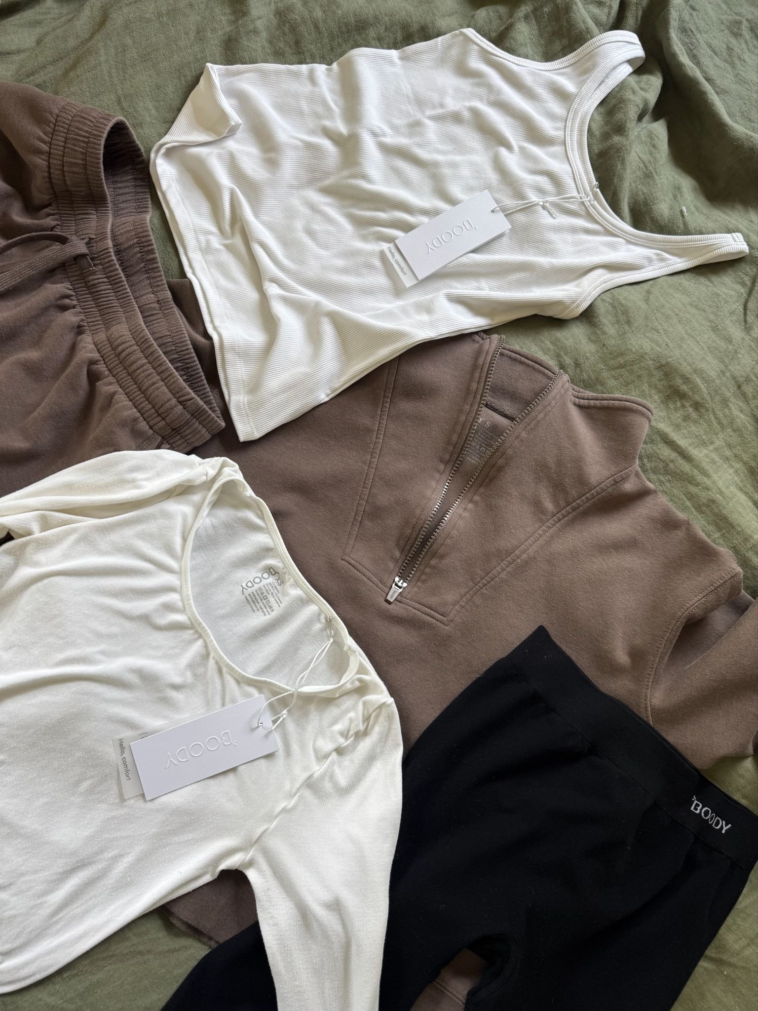 A neatly arranged flat lay of neutral-toned clothing items, including tops, shorts, and leggings, all with brand tags reading "Boody," displayed on a green fabric background.