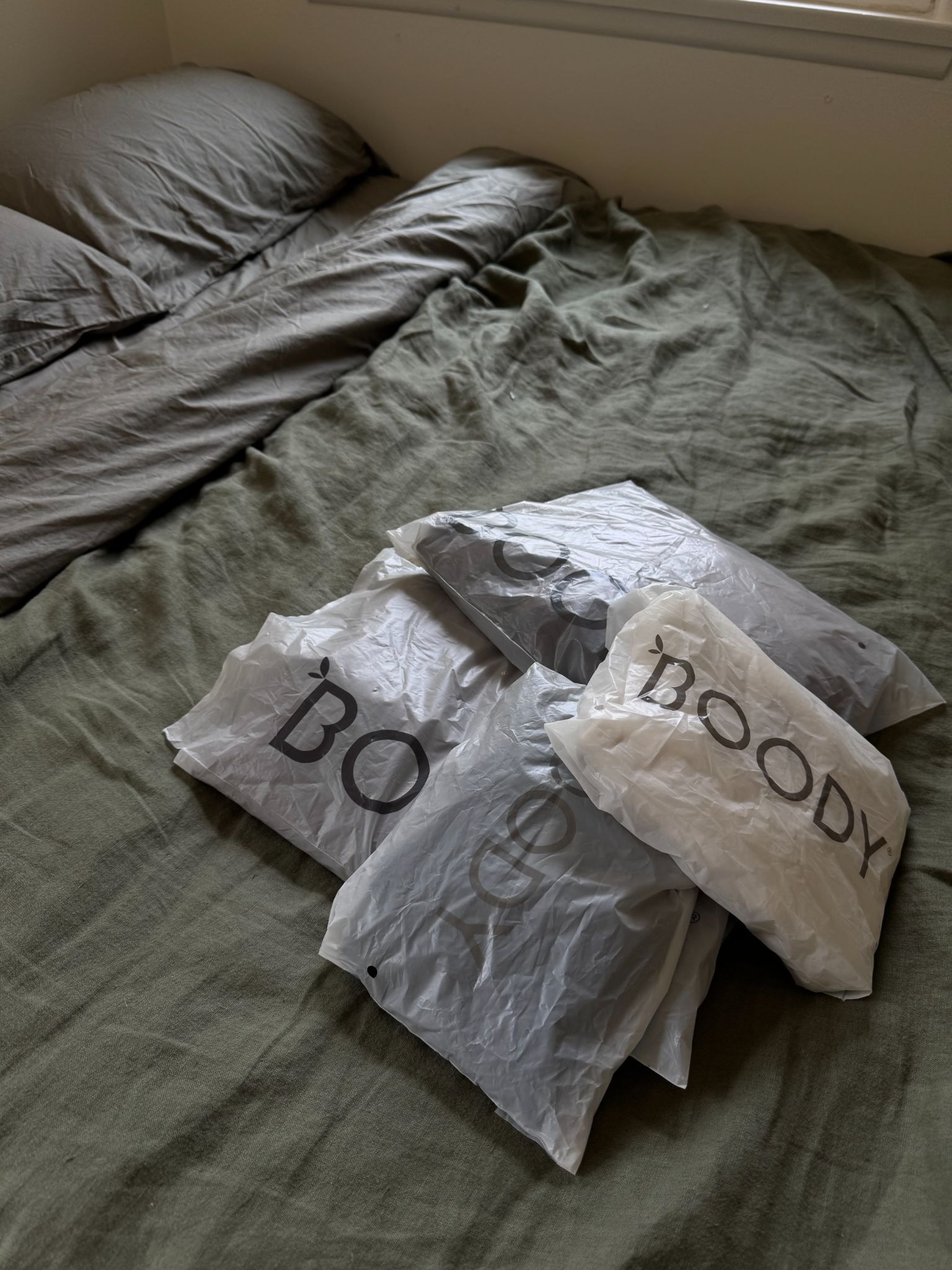 Four plastic-wrapped clothing packages labeled "BOODY" are placed on a green bedspread next to a neatly made gray pillow and blanket.