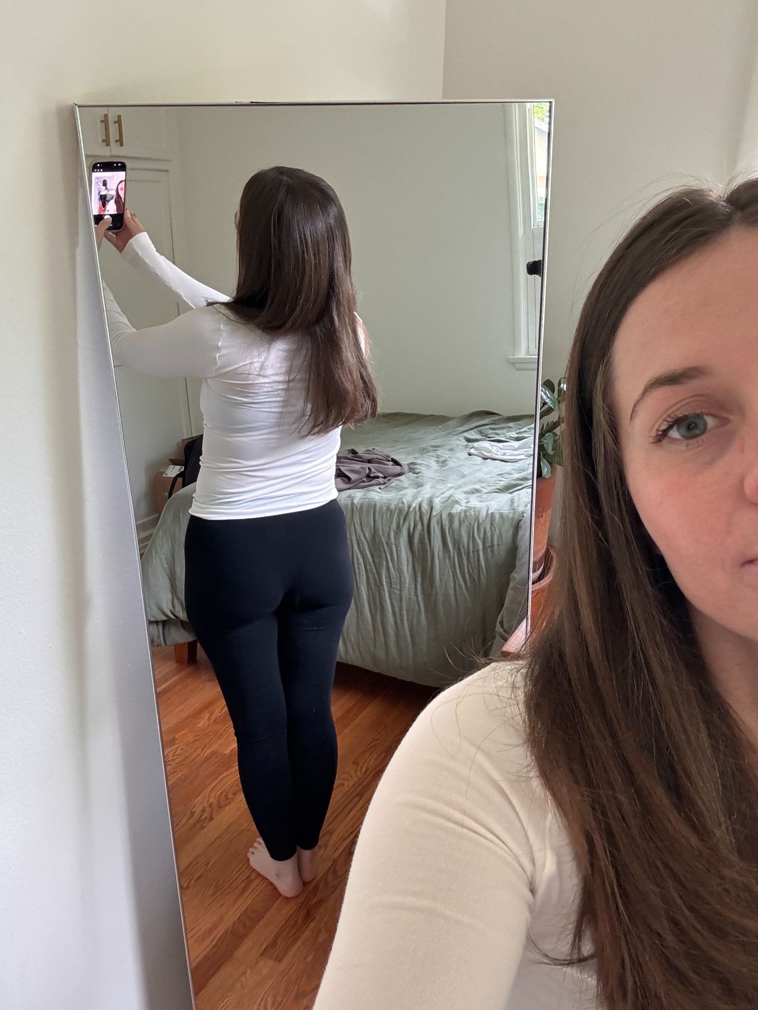 A woman stands in front of a mirror taking a photo, capturing both her reflection from behind and part of her face in the foreground. A bed and window are in the background.