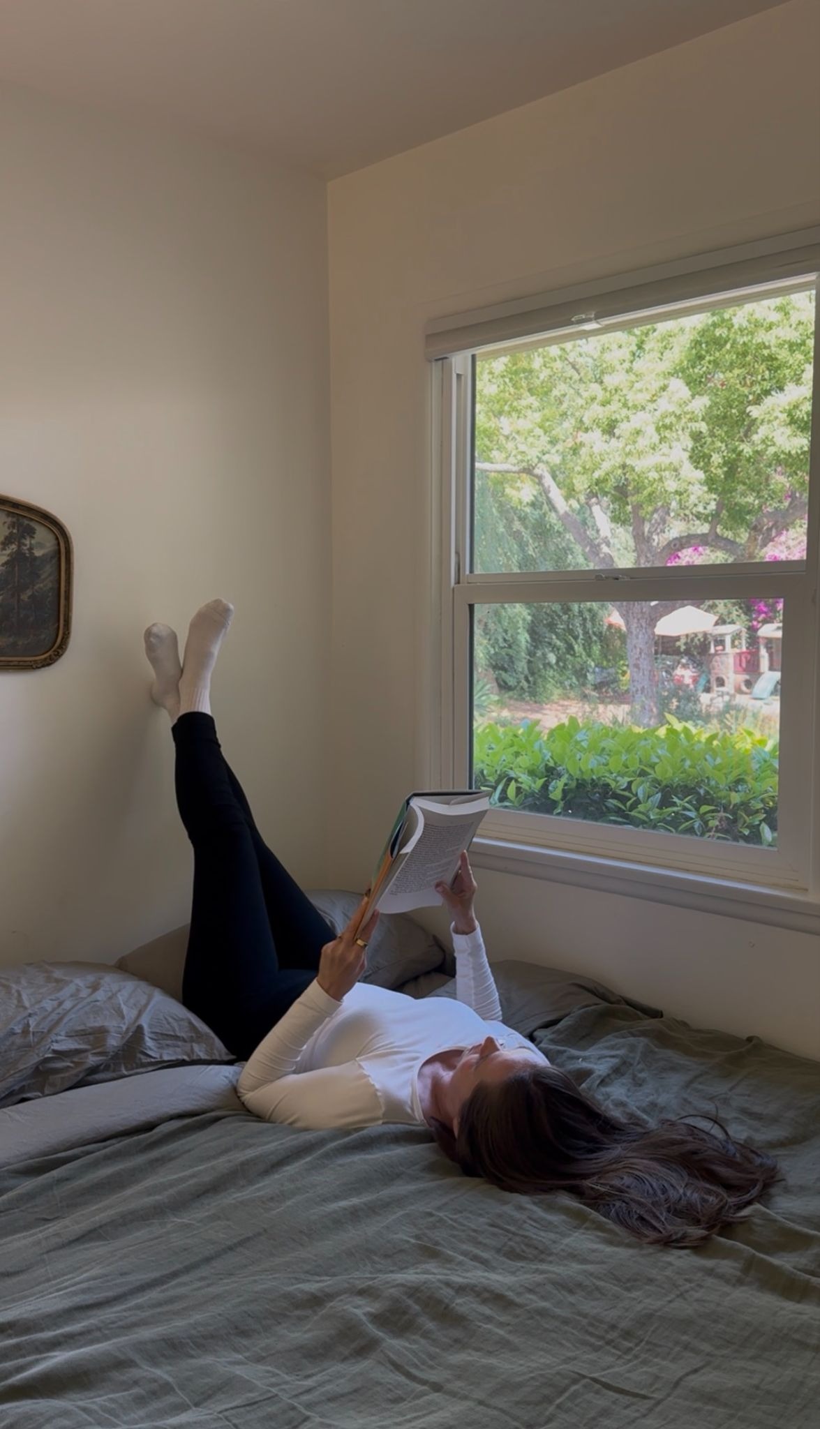 A person lies on a bed with their legs up against the wall, reading a book by an open window with trees visible outside.