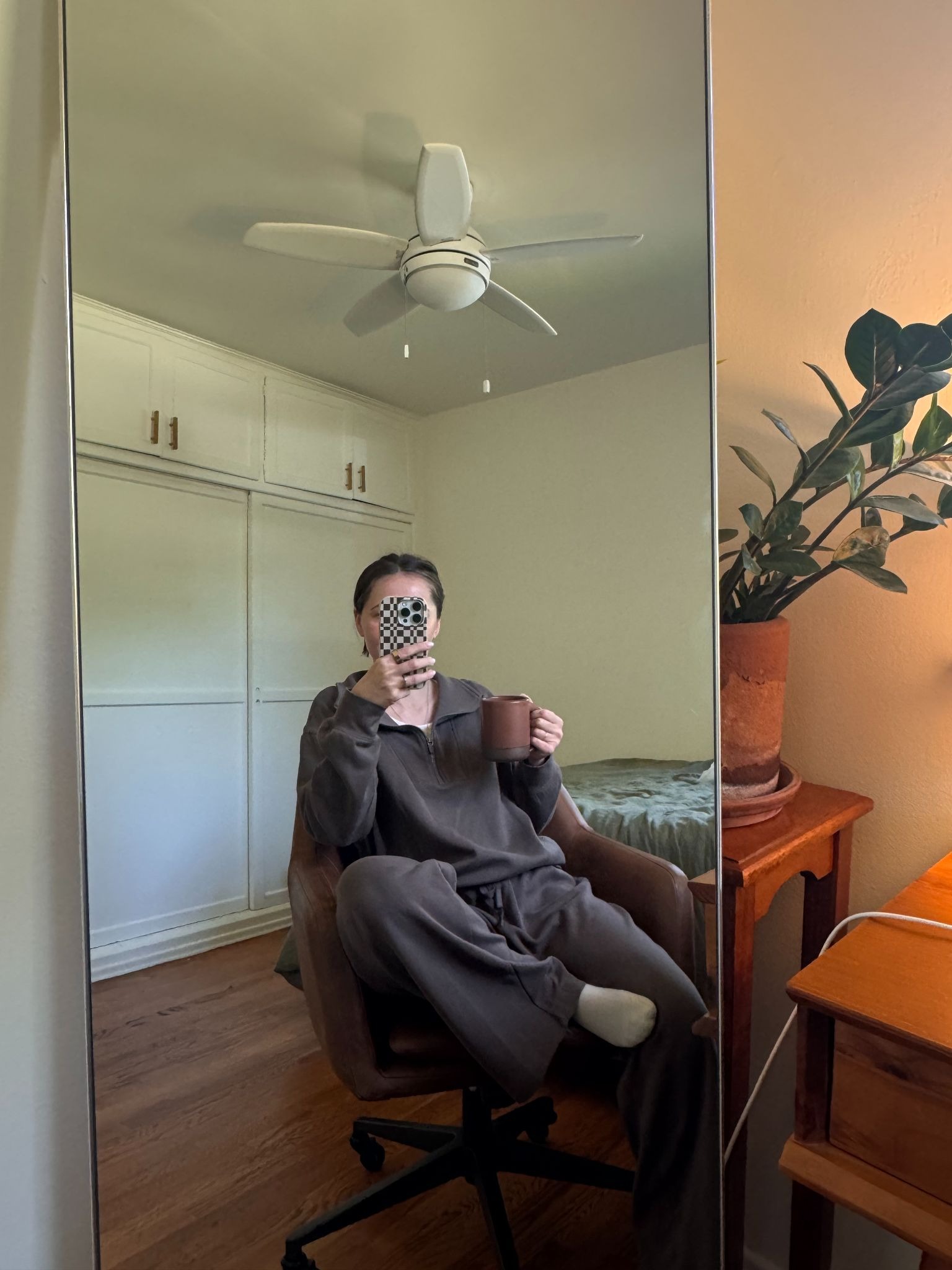 Person sitting on a chair in front of a mirror, holding a mug and a phone in a bedroom with white closets, a ceiling fan, and a potted plant on a wooden desk.